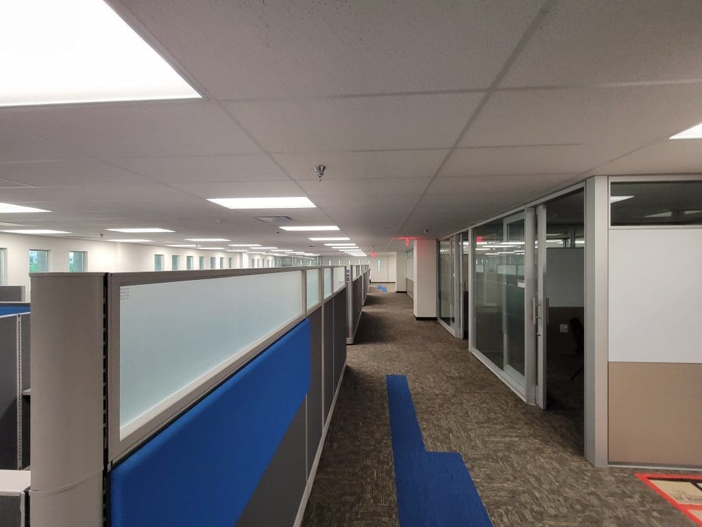 A modern office interior with cubicles on the left and glass-walled meeting rooms on the right, featuring neutral colors and overhead fluorescent lighting.