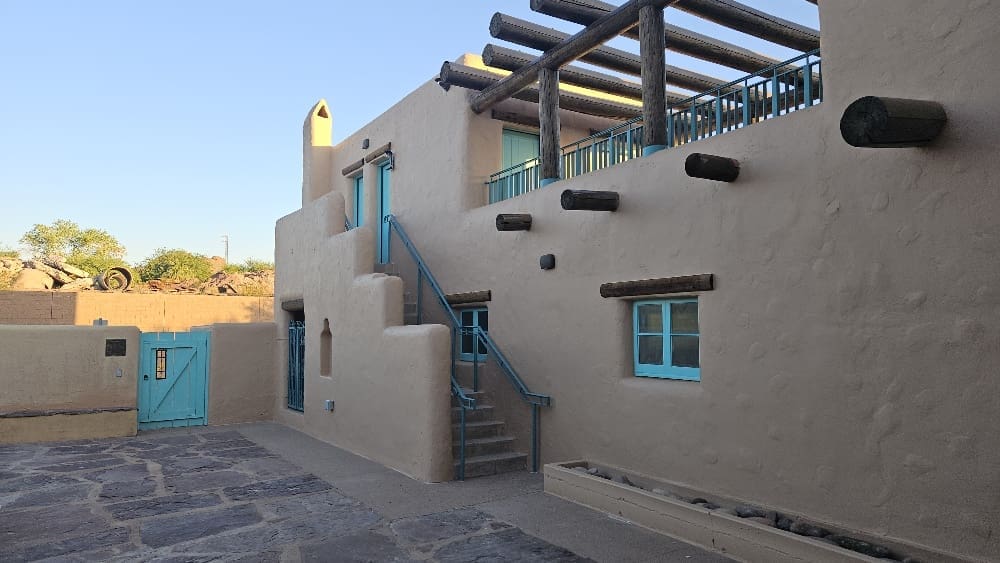 Adobe-style building with tan stucco walls, turquoise doors and windows, wooden beams, an exterior staircase, and a stone courtyard under a clear sky.
