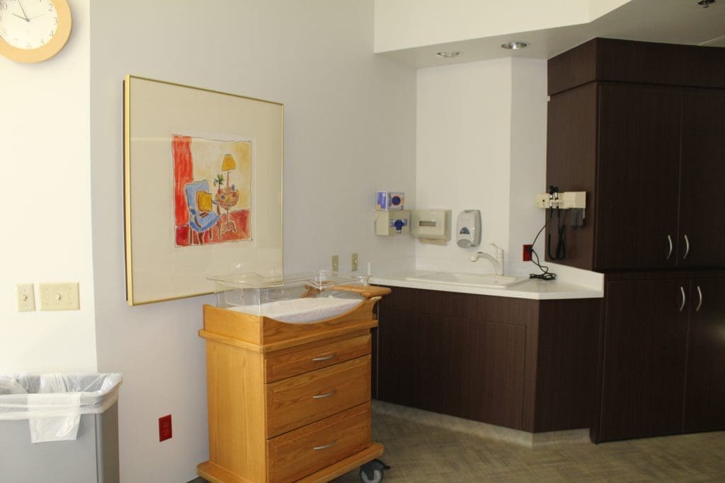 A hospital room corner with a wooden bassinet, sink, medical equipment on the wall, dark cabinets, a trash bin, and a framed artwork.