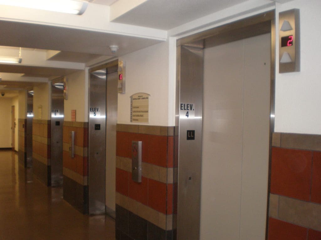 Four elevator doors are lined up in a hallway, each labeled with numbers and letters, with lit floor indicators above showing different floors.