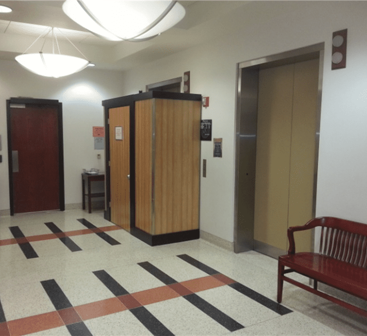 A hallway with an elevator, a wooden bench, a wooden phone booth, a closed door, and overhead lighting. The floor has a pattern of black and red stripes.