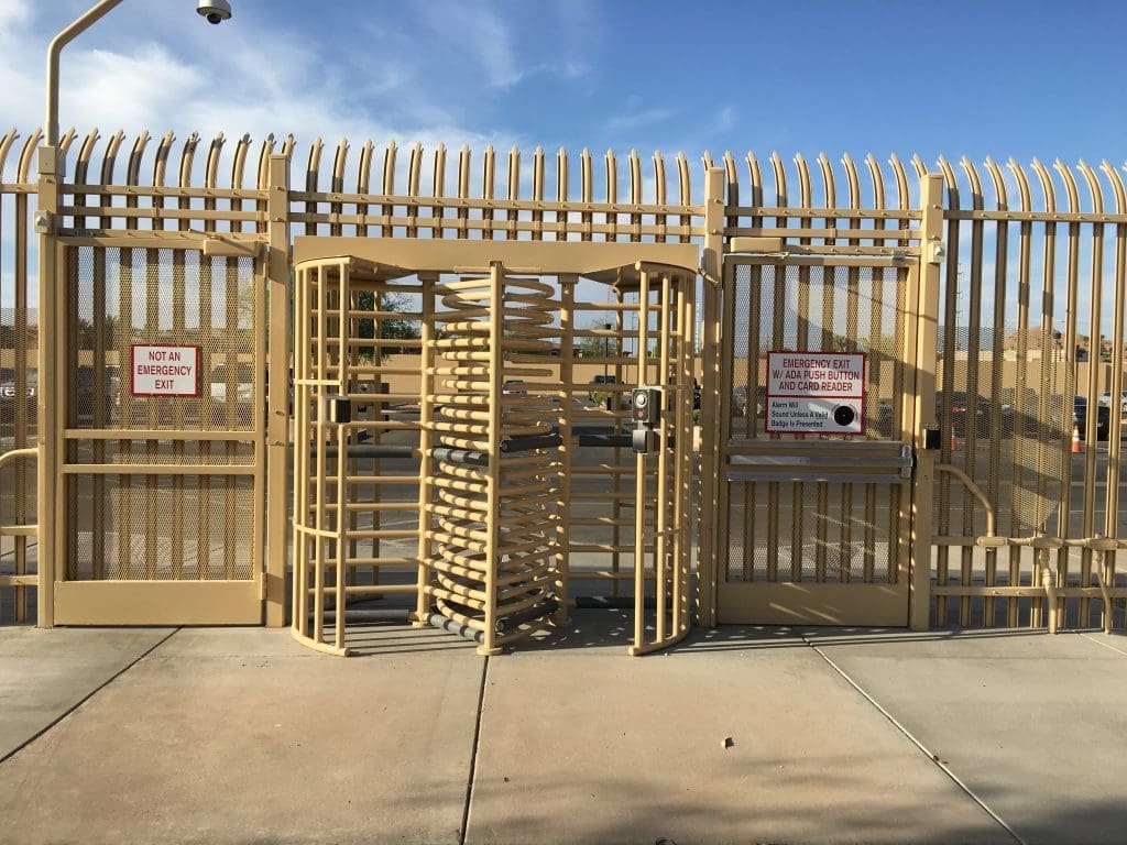 A tall metal security fence with a central revolving turnstile gate and warning signs stating "Not an Emergency Exit" and "Emergency Exit Can Be Used Only in an Emergency.