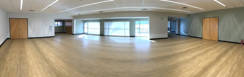 Spacious, empty room with wood flooring, white and light green walls, ceiling lights, four closed wooden doors, large windows, and a fire extinguisher in the corner.