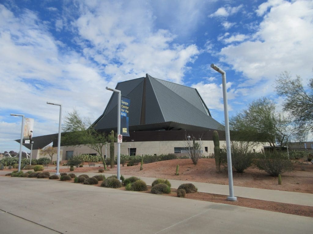 A modern building with a geometric dark roof, surrounded by desert plants and a sidewalk under a partly cloudy sky.
