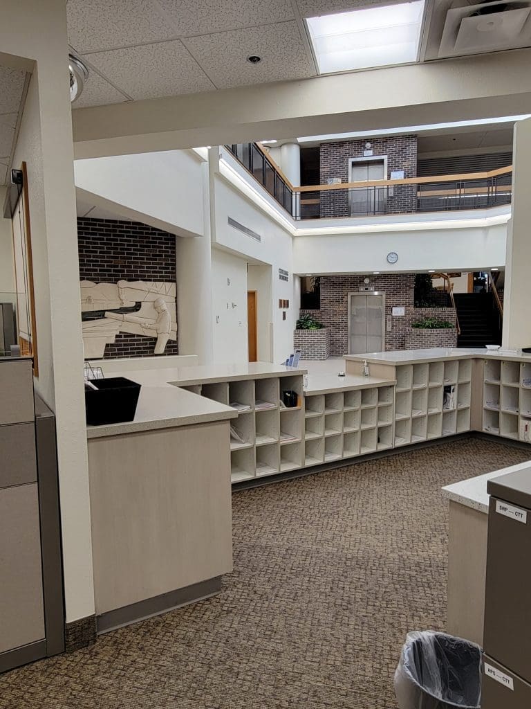 A reception area with open cubby shelves, a counter, and a view of a brick wall mural, an elevator, and a staircase in the background.