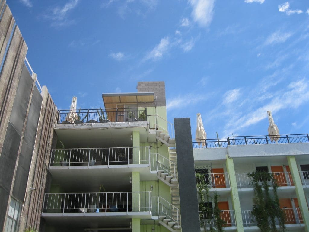 A multi-story building with green walls, white railings, and orange doors, under a blue sky with scattered clouds. Outdoor umbrellas are visible on the rooftop.