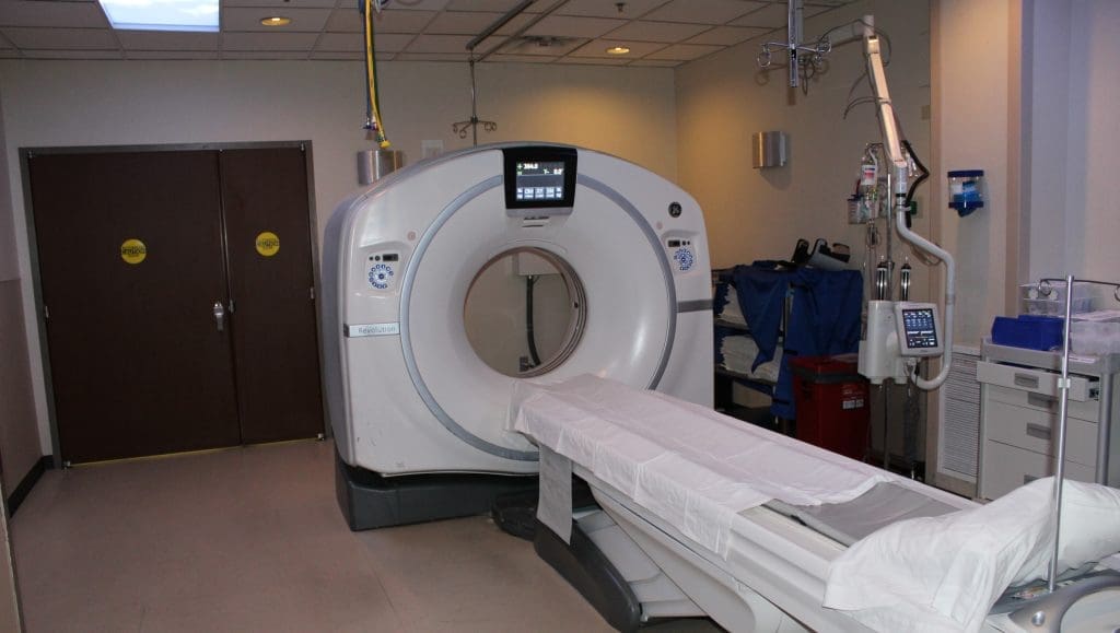 A CT scanner with a patient table is positioned in a hospital imaging room with medical equipment and closed double doors.