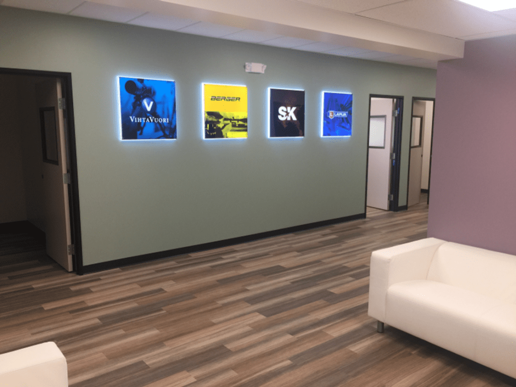 A hallway with four illuminated logo signs on a green wall, light wood flooring, white couches, and several open doorways.