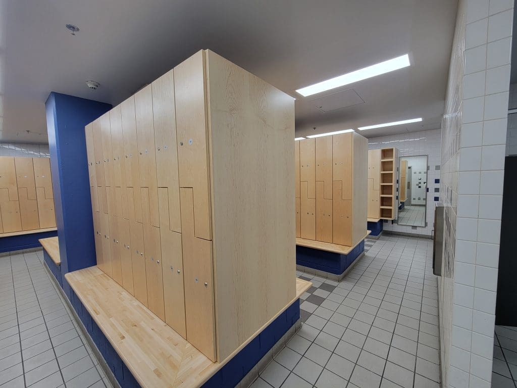 Locker room with light wood lockers, tiled floor, blue accent walls, benches, and mirrors in the background under fluorescent lighting.