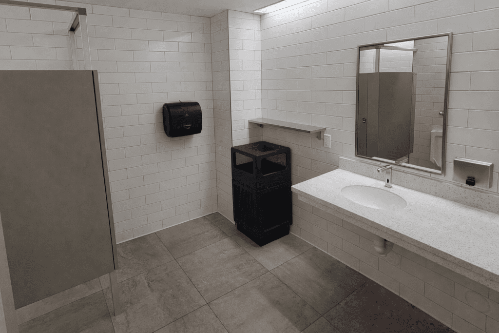 A public restroom with white tiled walls, a counter with a sink and mirror, a paper towel dispenser, a trash bin, and a partially visible stall.