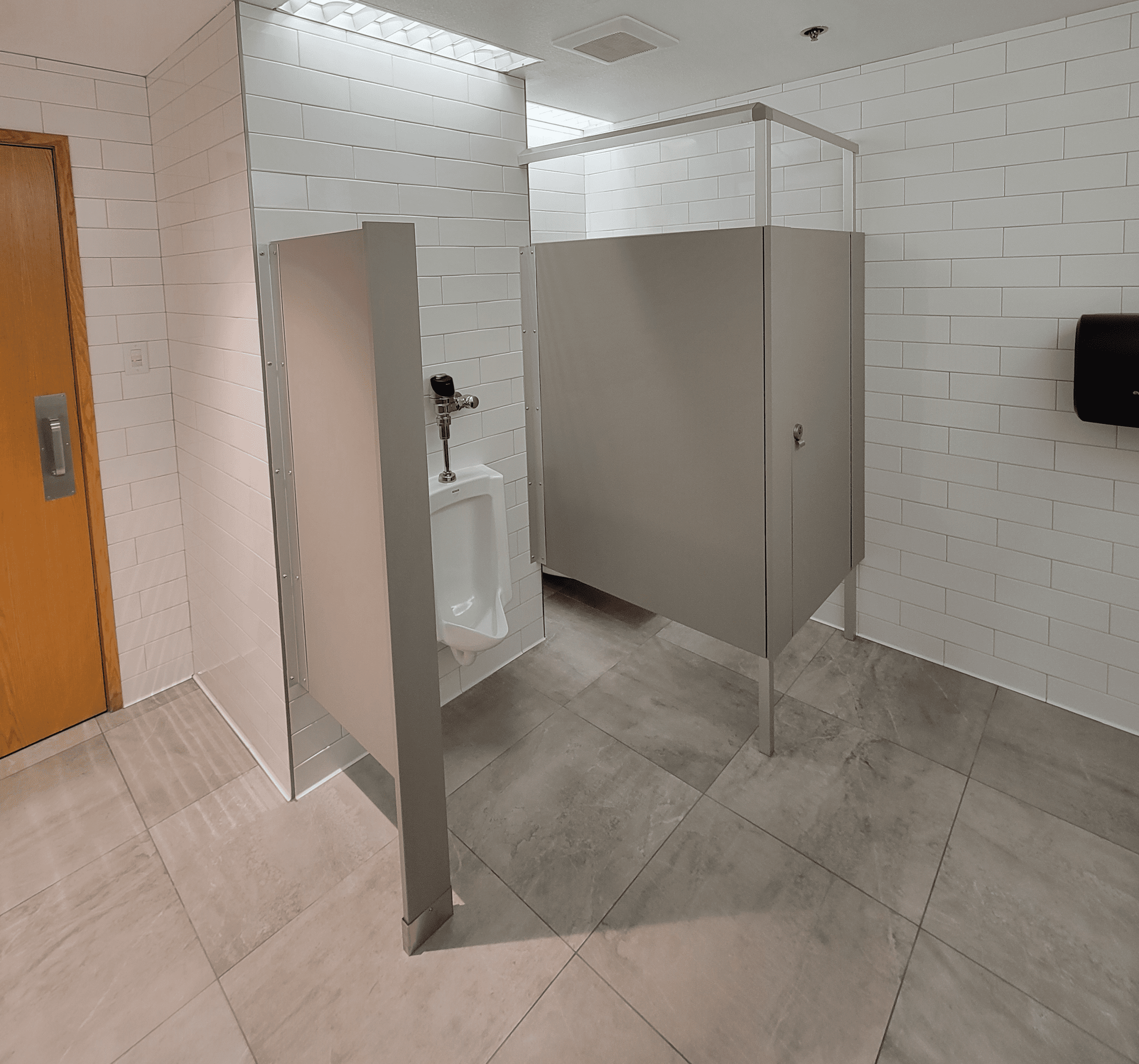 A public restroom with a urinal partially enclosed by a divider, a toilet stall with a closed door, white tiled walls, and a gray tiled floor.