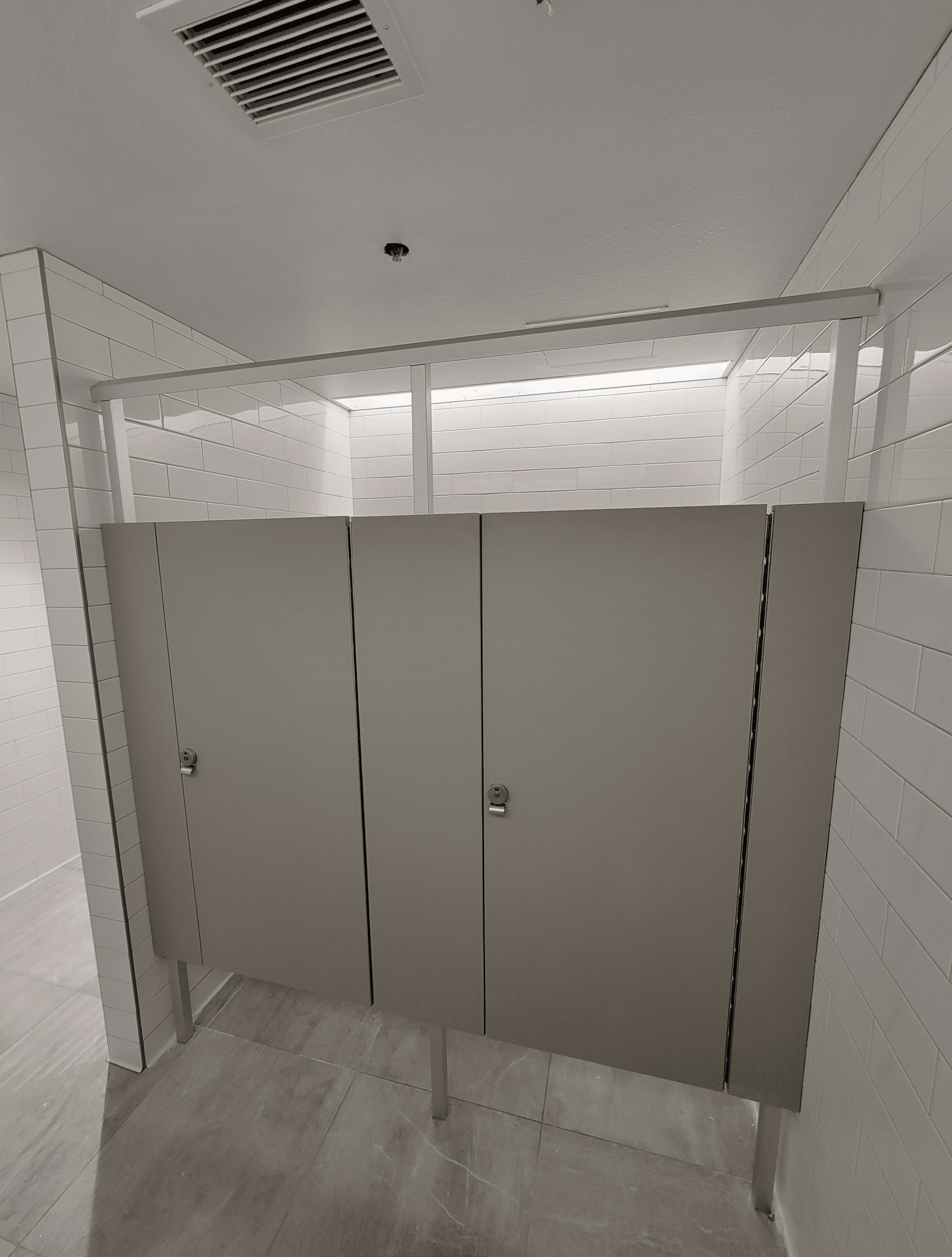 Two closed gray bathroom stall doors in a clean, modern restroom with white tiled walls and a tiled floor.
