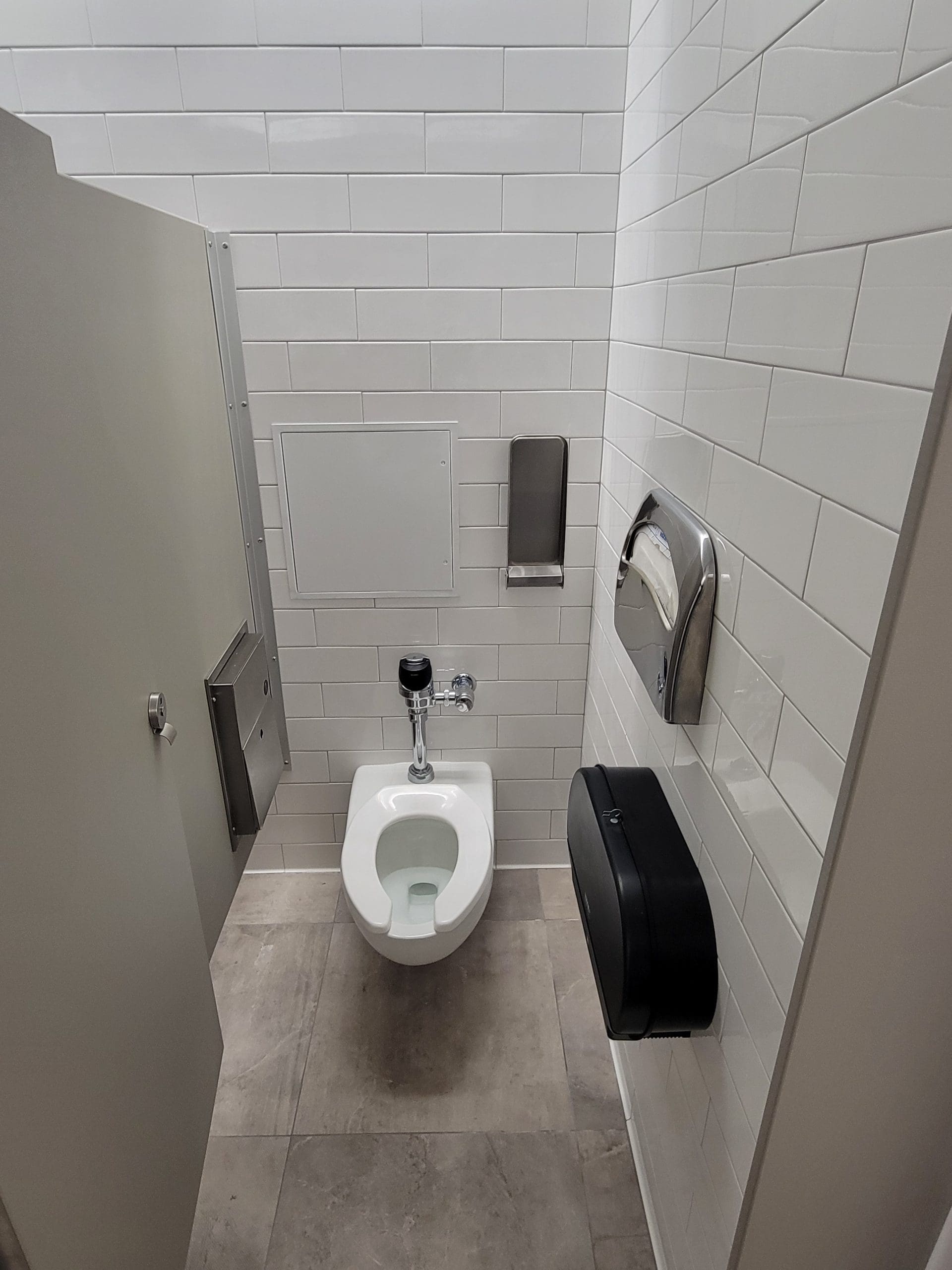 A public restroom stall with a white toilet, wall-mounted toilet paper dispenser, paper towel dispenser, and tiled walls and floor.