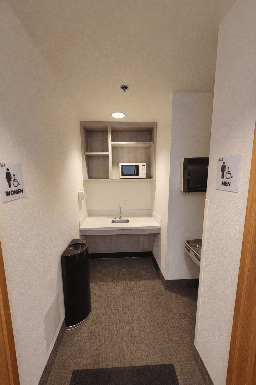 A small room with a sink, microwave, shelves, trash can, paper towel dispenser, and water fountain; signs for accessible men's and women's restrooms are on the walls.